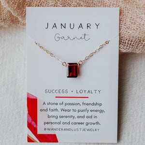 May include: A gold necklace with a red garnet gemstone pendant. The card reads "JANUARY Garnet SUCCESS + LOYALTY A stone of passion, friendship and faith. Wear to purify energy, bring serenity, and aid in personal and career growth. @WANDERANDLUST JEWELRY"