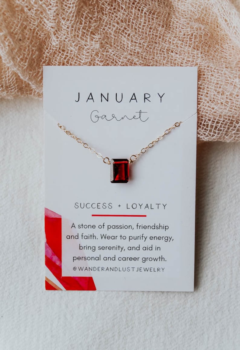 May include: A gold necklace with a red garnet gemstone pendant. The card reads "JANUARY Garnet SUCCESS + LOYALTY A stone of passion, friendship and faith. Wear to purify energy, bring serenity, and aid in personal and career growth. @WANDERANDLUST JEWELRY"