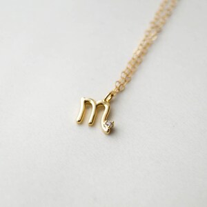 May include: A gold necklace with a small letter 'M' pendant featuring a single clear gemstone.