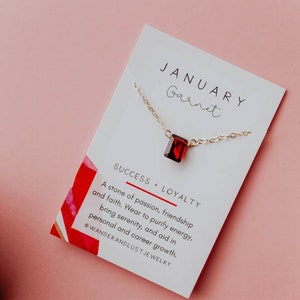 May include: A gold necklace with a red garnet pendant on a white card with the text "JANUARY Garnet SUCCESS LOYALTY A stone of passion, friendship and faith. Wear to purify energy, bring serenity, and aid in personal and career growth. @WANDERANDLUST JEWELRY"