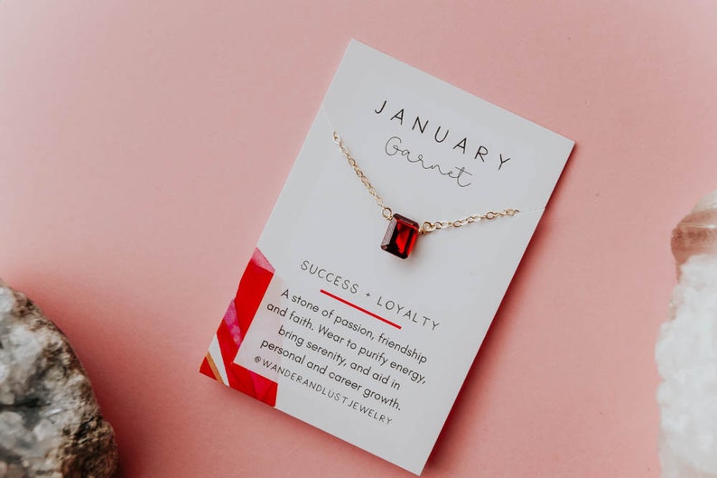 May include: A gold necklace with a red garnet pendant on a white card with the text "JANUARY Garnet SUCCESS LOYALTY A stone of passion, friendship and faith. Wear to purify energy, bring serenity, and aid in personal and career growth. @WANDERANDLUST JEWELRY"