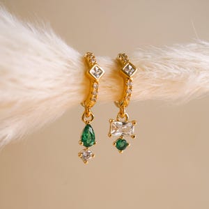 May include: A pair of gold hoop earrings featuring small clear stones along the band and a square stone at the top. One earring has a dangling pear-shaped green stone and a small clear stone, while the other has a rectangular clear stone and a round green stone.