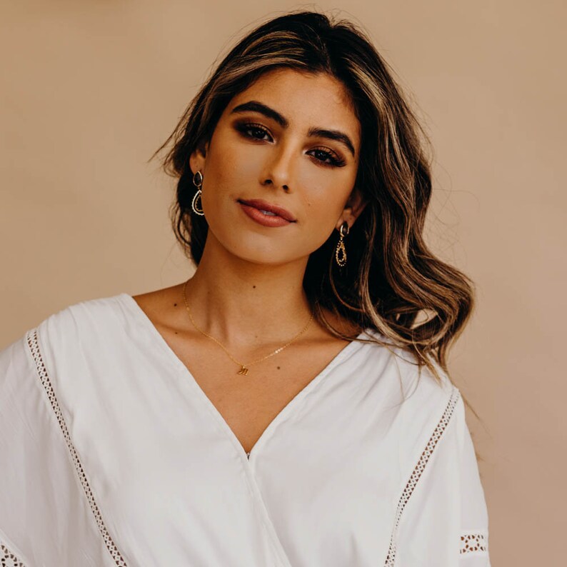 May include: A woman with long brown hair wearing a white blouse with lace trim. She is wearing gold earrings and a gold necklace with a small letter pendant.