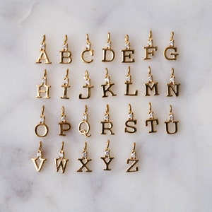 May include: A set of 26 gold-tone alphabet letter charms, each with a single clear rhinestone. The charms are arranged in alphabetical order on a white marble surface.