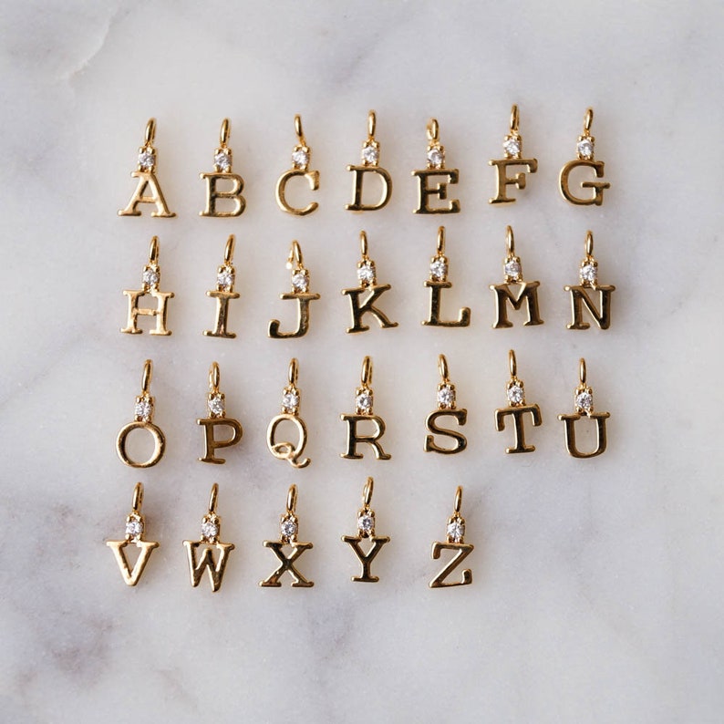 May include: A set of 26 gold-tone alphabet letter charms, each with a single clear rhinestone. The charms are arranged in alphabetical order on a white marble surface.