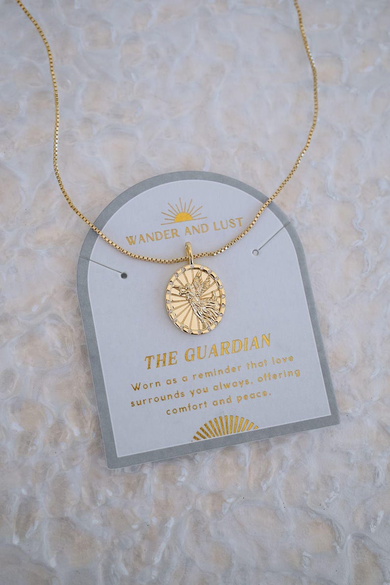 Puede incluir: Collar con colgante dorado con un dije ovalado con un dise&ntilde;o de p&aacute;jaro. El dije cuelga de una cadena delicada. El collar descansa sobre una tarjeta con el texto "WANDER AND LUST" y "THE GUARDIAN".