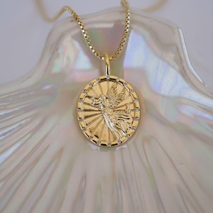 Puede incluir: Collar con colgante dorado con un dije circular detallado con un dise&ntilde;o de figura alada. El dije est&aacute; adornado con peque&ntilde;as gemas transparentes alrededor del borde y cuelga de una delicada cadena. El collar se exhibe sobre una superficie nacarada.