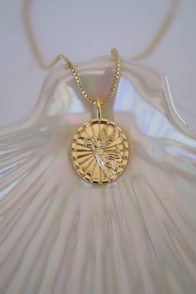Puede incluir: Collar con colgante dorado con un dije circular detallado con un dise&ntilde;o de figura alada. El dije est&aacute; adornado con peque&ntilde;as gemas transparentes alrededor del borde y cuelga de una delicada cadena. El collar se exhibe sobre una superficie nacarada.