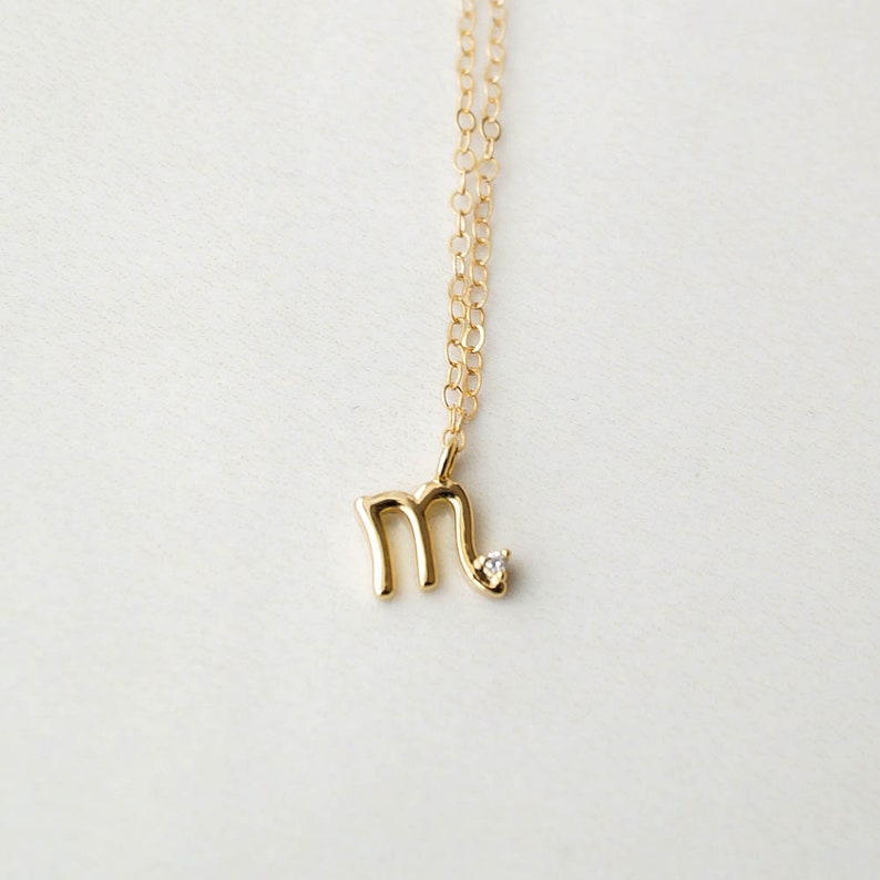 May include: A gold chain necklace with a small gold pendant in the shape of the letter "M" with a single diamond accent.