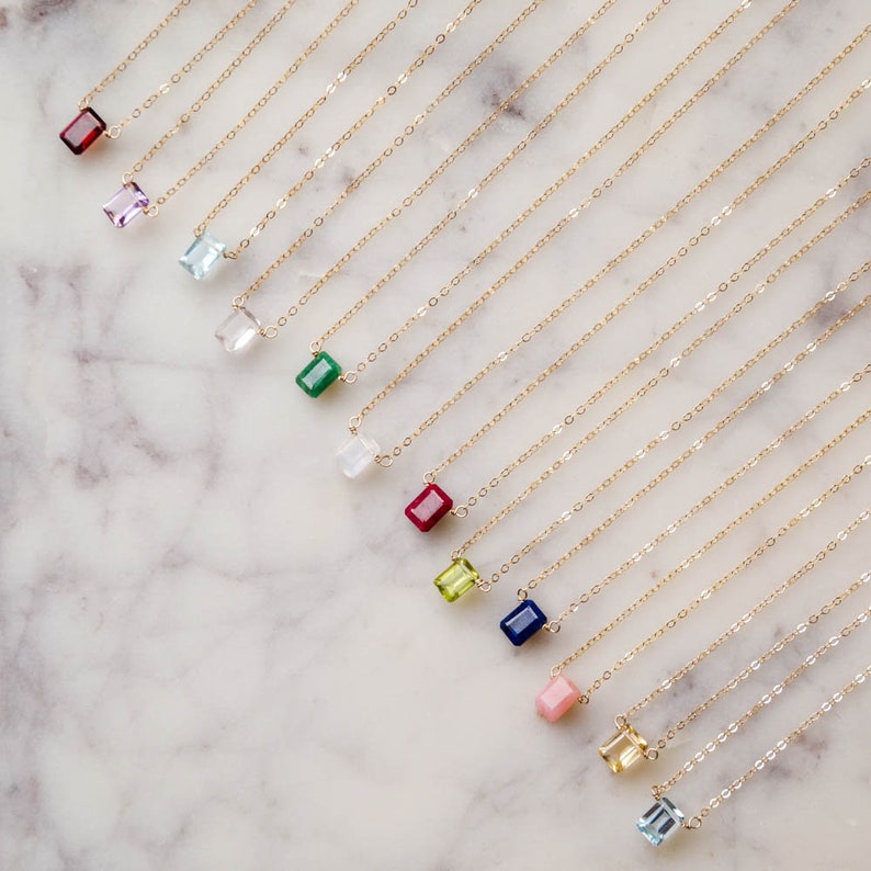 May include: A collection of delicate gold necklaces with various colored gemstone pendants, including red, pink, green, blue, and clear. The necklaces are arranged in a row on a white marble surface.