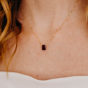 May include: A delicate gold chain necklace with a small, square-shaped, dark red gemstone pendant.