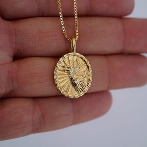 Puede incluir: Collar con colgante dorado con un dije circular detallado con un dise&ntilde;o de &aacute;ngel. El dije est&aacute; adornado con peque&ntilde;as piedras transparentes alrededor del borde y cuelga de una delicada cadena dorada. El collar se muestra sobre un fondo neutro.
