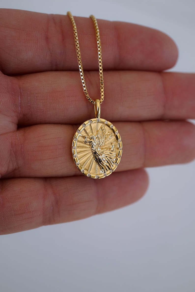Puede incluir: Collar con colgante dorado con un dije circular detallado con un dise&ntilde;o de &aacute;ngel. El dije est&aacute; adornado con peque&ntilde;as piedras transparentes alrededor del borde y cuelga de una delicada cadena dorada. El collar se muestra sobre un fondo neutro.