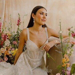 May include: A woman wearing a white, beaded, sleeveless gown with a sweetheart neckline. She is sitting in front of a floral arrangement with pink, orange, and yellow flowers.