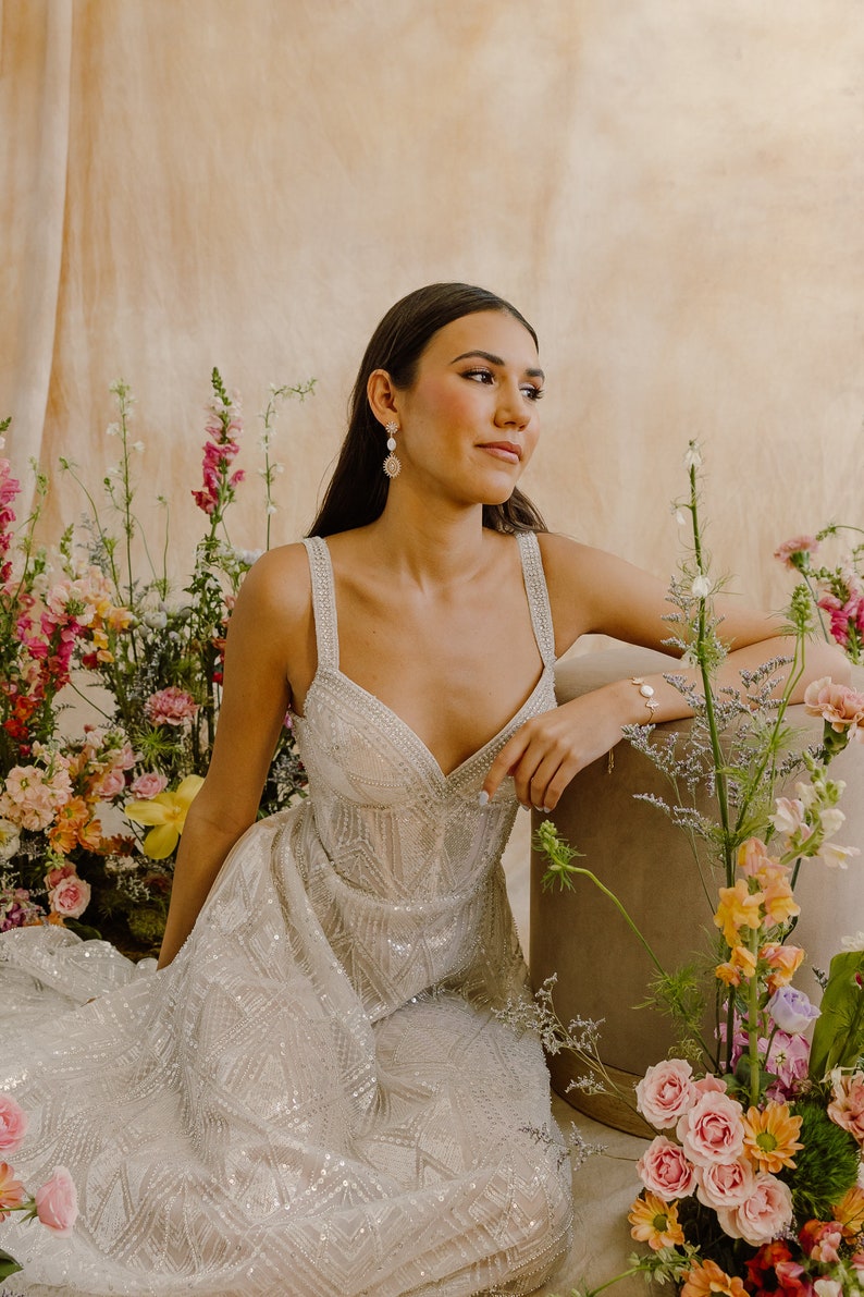 May include: A woman wearing a white, beaded, sleeveless gown with a sweetheart neckline. She is sitting in front of a floral arrangement with pink, orange, and yellow flowers.