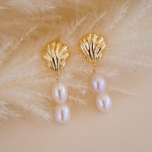 Op de afbeelding: Goudkleurige oorbellen in de vorm van een zeeschelp met twee bungelende witte parels. De oorbellen zijn geplaatst tegen een zachte, neutrale achtergrond met veervormige accenten, wat een elegante en natuurlijke uitstraling creëert. Een stijlvol accessoire.
