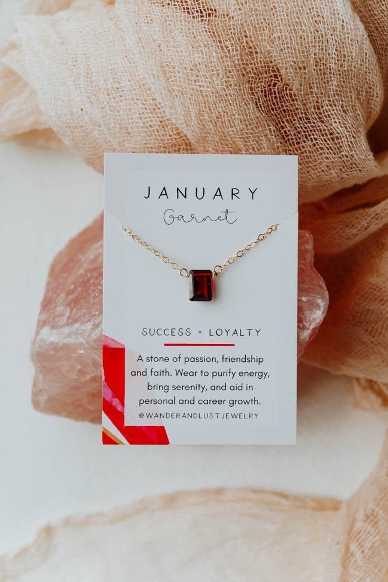 May include: A white card with the text "JANUARY Garnet" and "SUCCESS + LOYALTY" printed on it. A gold chain necklace with a red garnet gemstone pendant is displayed on the card. The text below the gemstone reads "A stone of passion, friendship and faith. Wear to purify energy, bring serenity, and aid in personal and career growth. @WANDERANDLUST JEWELRY"