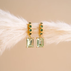 May include: A pair of gold hoop earrings featuring small, round, dark green stones set along the hoop and a single, rectangular, light green emerald-cut stone dangling from each earring. The metal is polished gold, and the design is symmetrical.