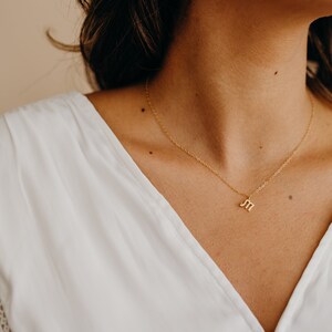 May include: A gold chain necklace with a small letter "M" pendant. The necklace is worn by a person with long brown hair and a white shirt.