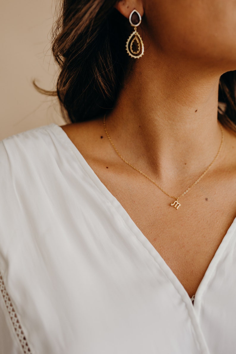 May include: A gold chain necklace with a small letter "M" pendant. The necklace is worn by a person with long brown hair and a white shirt.