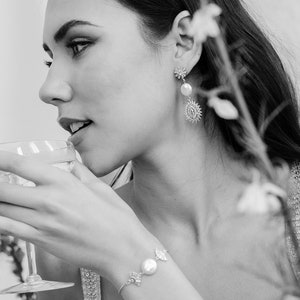 May include: A black and white photo of a person wearing a pearl and flower charm bracelet and earrings. The person is holding a glass of liquid and looking to the side.