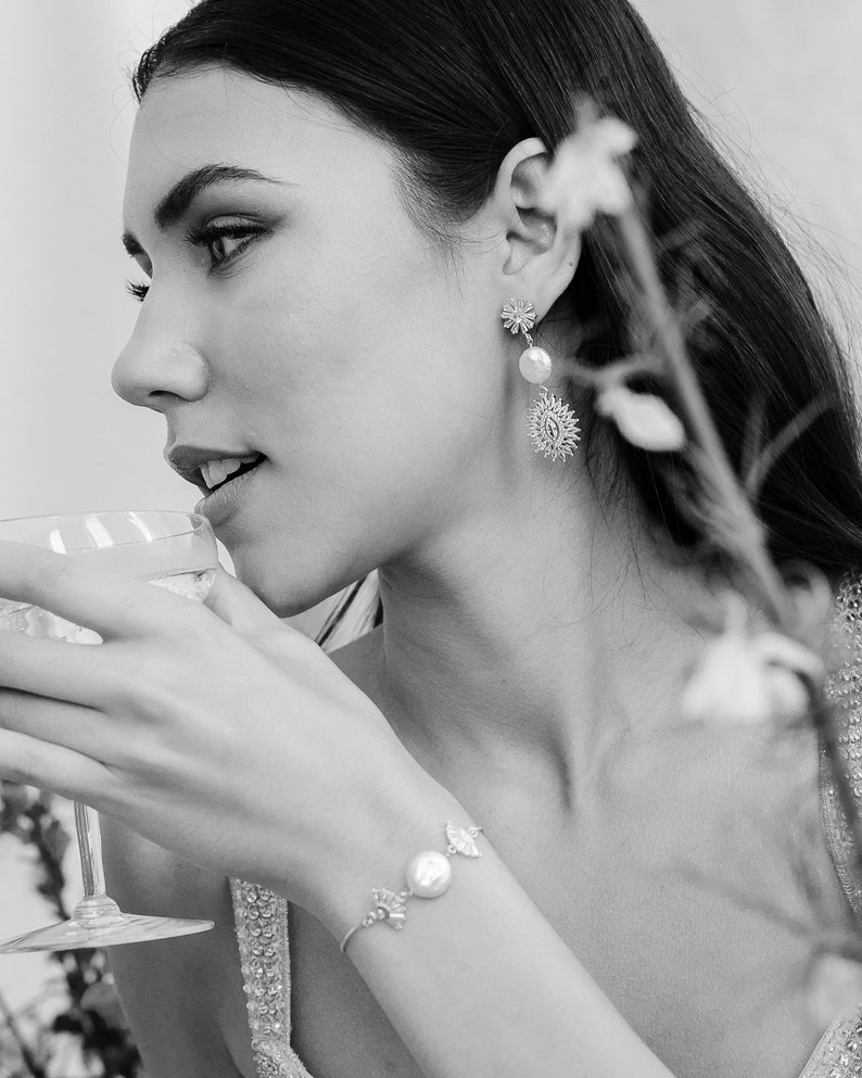 May include: A black and white photo of a person wearing a pearl and flower charm bracelet and earrings. The person is holding a glass of liquid and looking to the side.