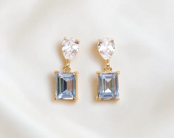 May include: A pair of gold-tone drop earrings featuring a clear, pear-shaped faceted stone at the top connected to a rectangular, light blue faceted stone at the bottom, both set in gold-coloured metal prongs with a secure hook attachment.