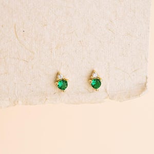 May include: A pair of gold stud earrings, each featuring a round, vibrant green gemstone set in a gold prong setting, topped with a single, smaller, clear white round gemstone, creating a delicate and balanced vertical design.