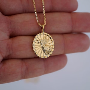 Puede incluir: Un collar con colgante dorado con un dise&ntilde;o detallado de una figura alada dentro de un marco circular. El colgante est&aacute; adornado con peque&ntilde;os acentos brillantes y cuelga de una delicada cadena. El collar es una joya.