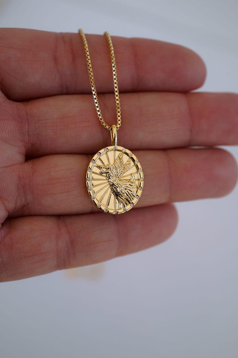 Puede incluir: Un collar con colgante dorado con un dise&ntilde;o detallado de una figura alada dentro de un marco circular. El colgante est&aacute; adornado con peque&ntilde;os acentos brillantes y cuelga de una delicada cadena. El collar es una joya.