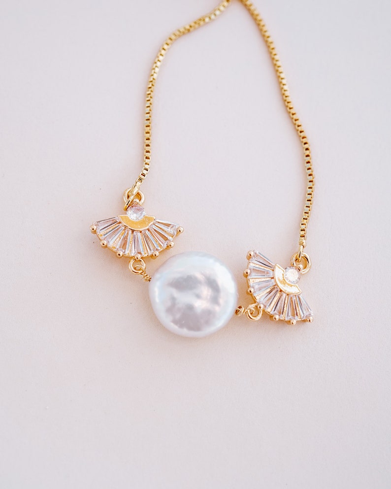 May include: A delicate gold chain necklace with a large white pearl and two gold fan-shaped charms adorned with clear rhinestones.