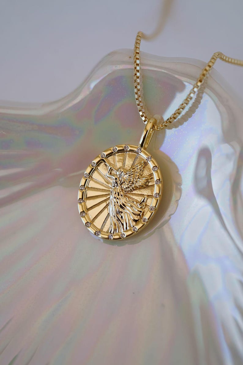 Puede incluir: Collar con colgante dorado con un relieve detallado de una figura alada dentro de un marco circular. El colgante est&aacute; adornado con peque&ntilde;as gemas transparentes alrededor del borde y cuelga de una delicada cadena. El collar se exhibe sobre una superficie nacarada.
