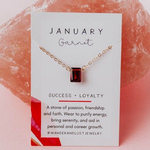 May include: A gold chain necklace with a red garnet gemstone pendant. The card reads "JANUARY Garnet SUCCESS + LOYALTY A stone of passion, friendship and faith. Wear to purify energy, bring serenity, and aid in personal and career growth. @WANDERANDLUST JEWELRY"