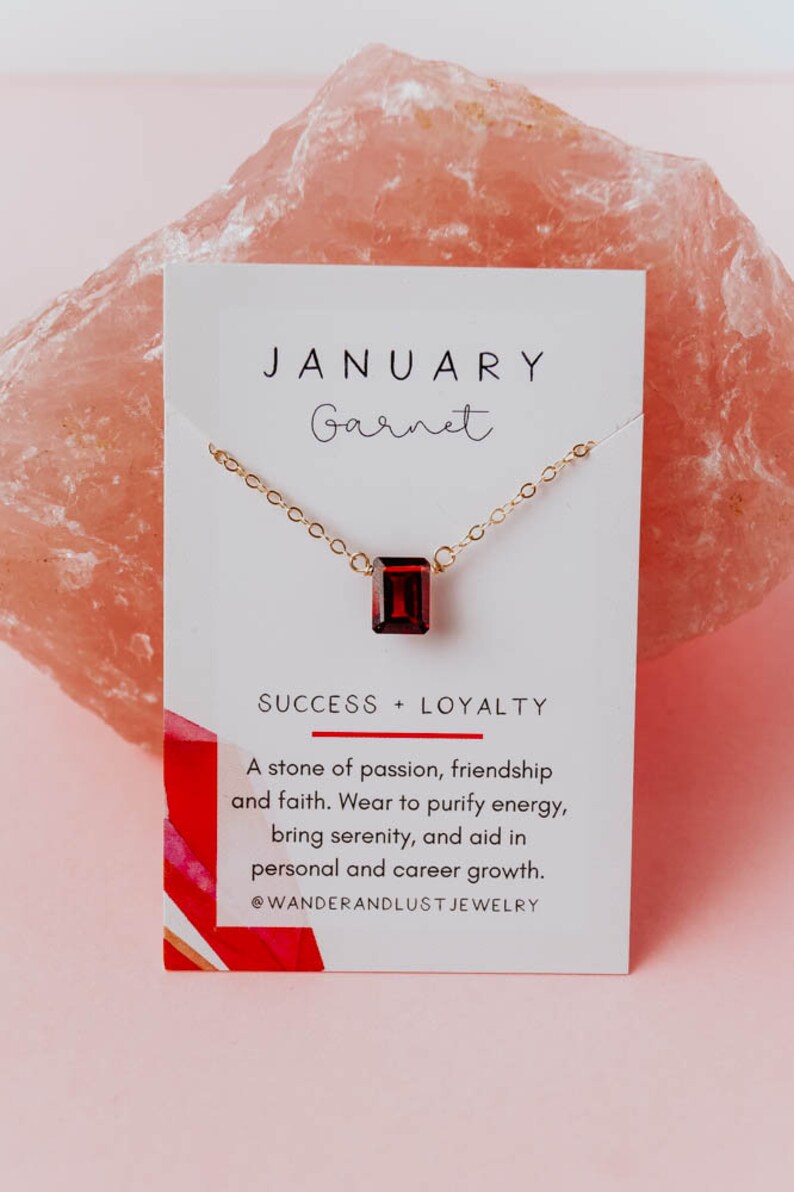 May include: A gold chain necklace with a red garnet gemstone pendant. The card reads "JANUARY Garnet SUCCESS + LOYALTY A stone of passion, friendship and faith. Wear to purify energy, bring serenity, and aid in personal and career growth. @WANDERANDLUST JEWELRY"