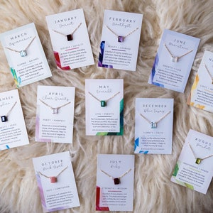 May include: Twelve cards with necklaces and birthstones for each month of the year. Each card has a different colored gemstone and a description of the stone's meaning. The cards are arranged in a circle on a white fluffy surface.
