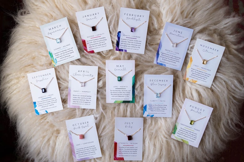 May include: Twelve cards with necklaces and birthstones for each month of the year. Each card has a different colored gemstone and a description of the stone's meaning. The cards are arranged in a circle on a white fluffy surface.
