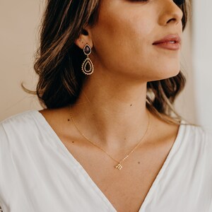 May include: A woman wearing a white top with a gold necklace featuring a small letter "M" pendant. She is wearing dangling earrings with a dark purple gemstone and gold accents.