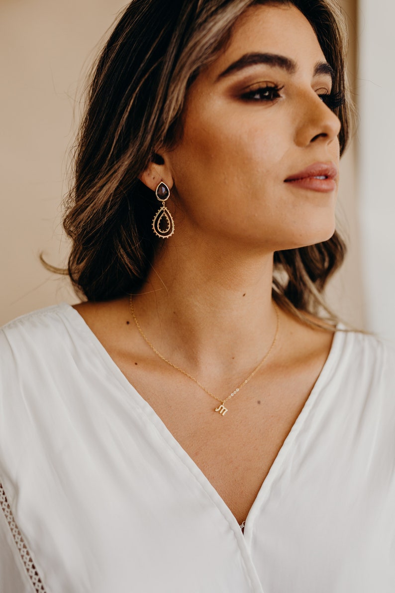 May include: A woman wearing a white top with a gold necklace featuring a small letter "M" pendant. She is wearing dangling earrings with a dark purple gemstone and gold accents.