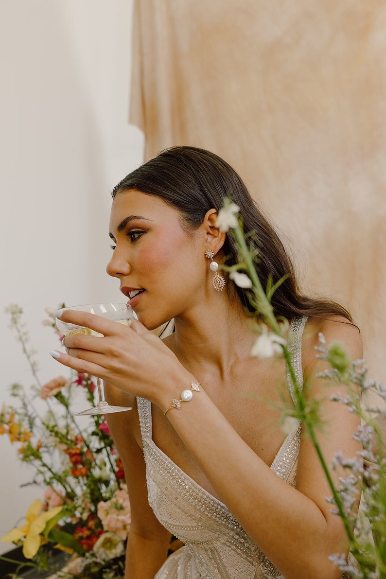 May include: A woman wearing a white beaded dress with a gold butterfly bracelet and pearl earrings. She is holding a glass of champagne and is surrounded by flowers.