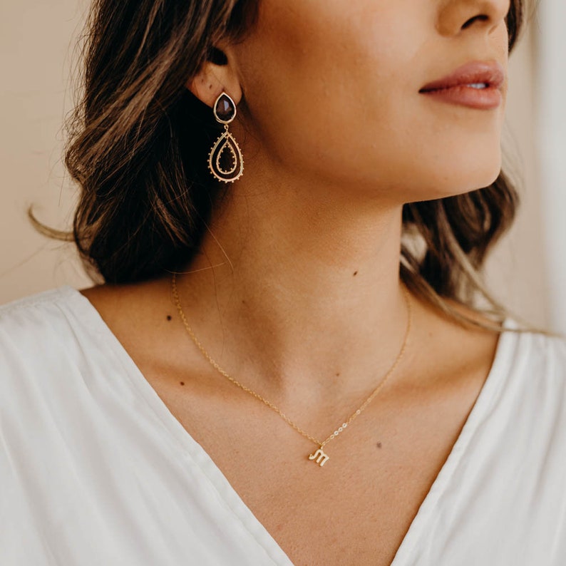 May include: A woman wearing a white shirt with a gold necklace featuring the initial "M". She is also wearing gold earrings with a dark purple stone.
