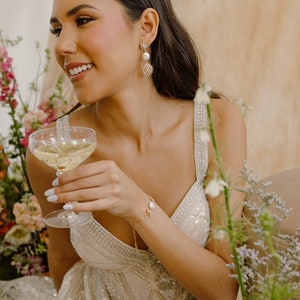 May include: A woman wearing a beaded, sequined, white dress with a gold chain necklace and a gold bracelet with a pearl accent. She is holding a champagne flute and wearing gold earrings with a pearl and a starburst design.