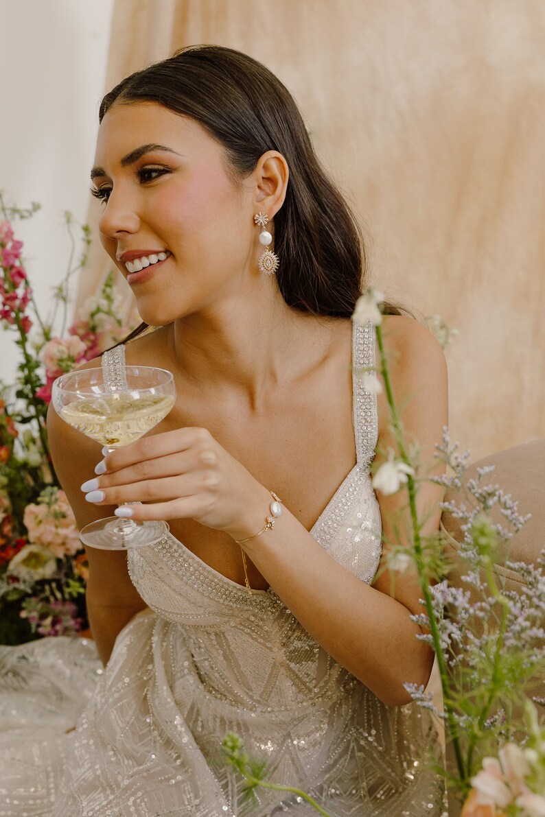 May include: A woman wearing a beaded, sequined, white dress with a gold chain necklace and a gold bracelet with a pearl accent. She is holding a champagne flute and wearing gold earrings with a pearl and a starburst design.