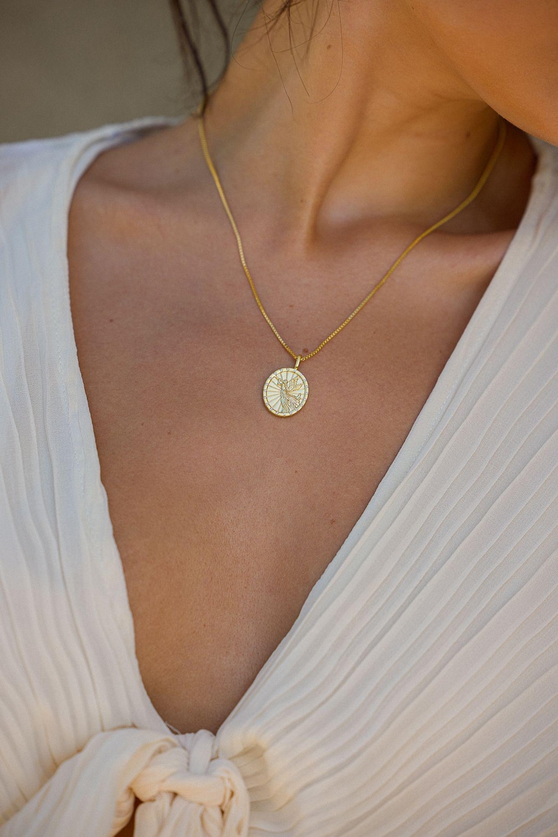 Puede incluir: Un collar dorado con un colgante circular con un dise&ntilde;o detallado. El colgante cuelga de una cadena fina. La persona lleva una blusa blanca plisada.