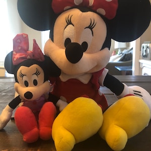 May include: Two plush Mickey Mouse characters, one large and one small. The larger plush is red and white with black ears and yellow feet. The smaller plush is pink and white with black ears and red feet. Both characters have black eyes and a black nose.