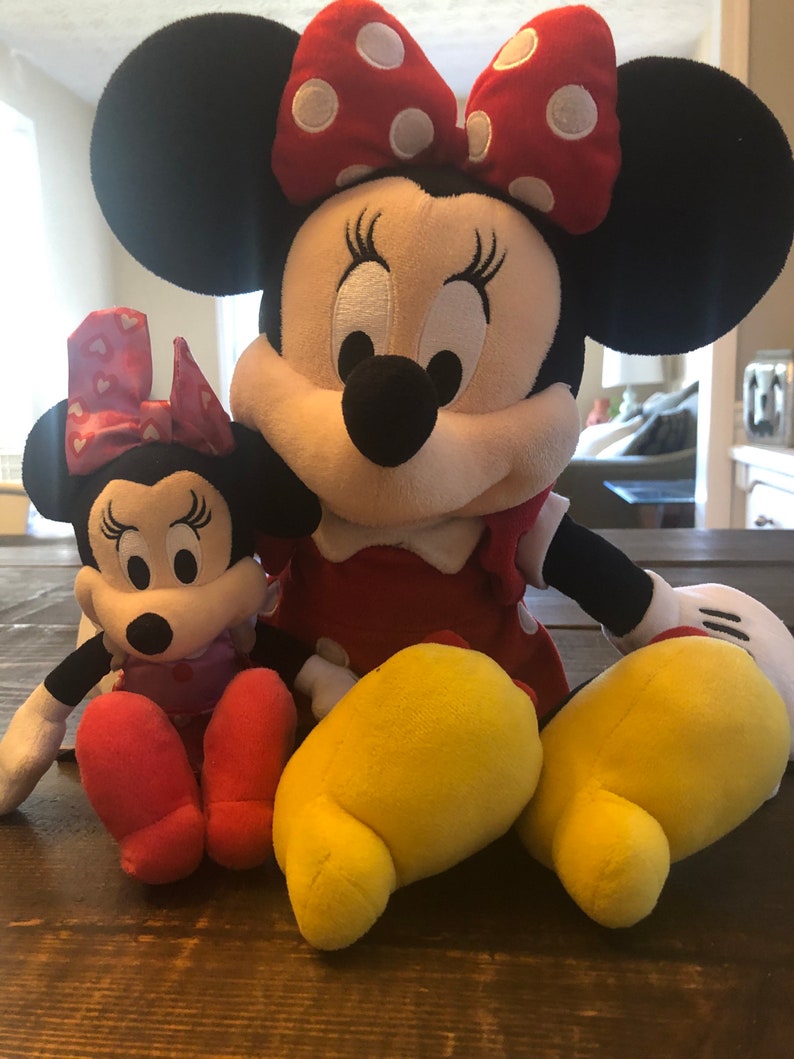 May include: Two plush Mickey Mouse characters, one large and one small. The larger plush is red and white with black ears and yellow feet. The smaller plush is pink and white with black ears and red feet. Both characters have black eyes and a black nose.