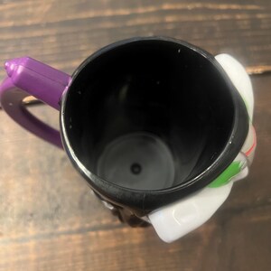 May include: Black ceramic mug with a purple handle. The mug features a white and green design on the side. The mug is empty and sits on a wooden surface.
