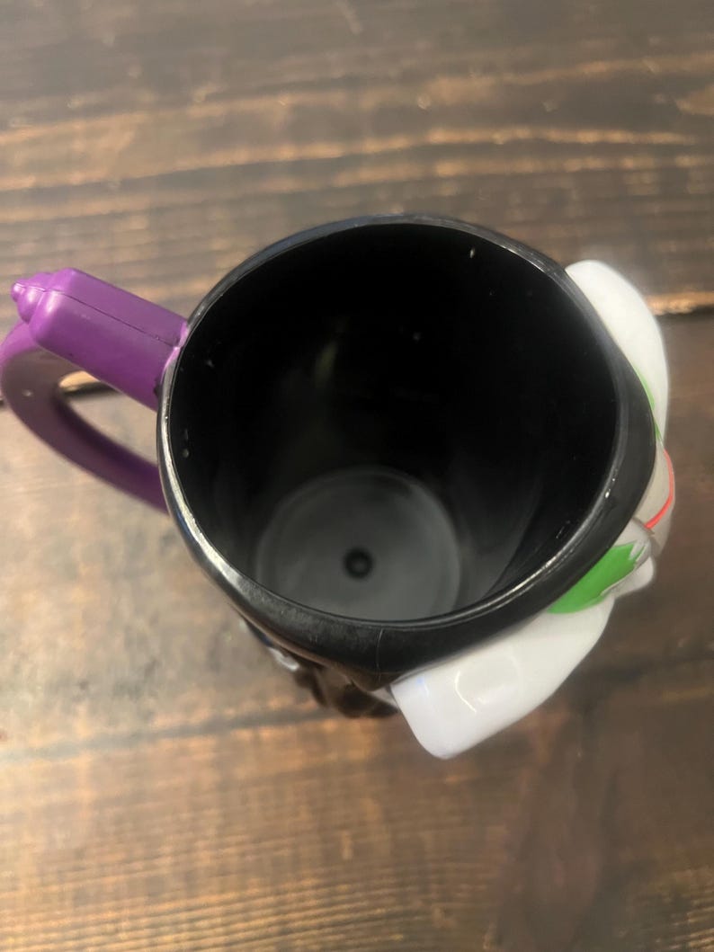 May include: Black ceramic mug with a purple handle. The mug features a white and green design on the side. The mug is empty and sits on a wooden surface.