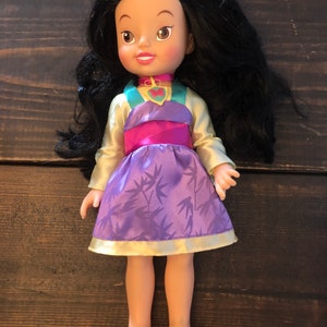 May include: A small doll with long black hair, wearing a purple and gold dress with a bamboo print. The doll has a pink and green top and a heart-shaped pendant necklace.