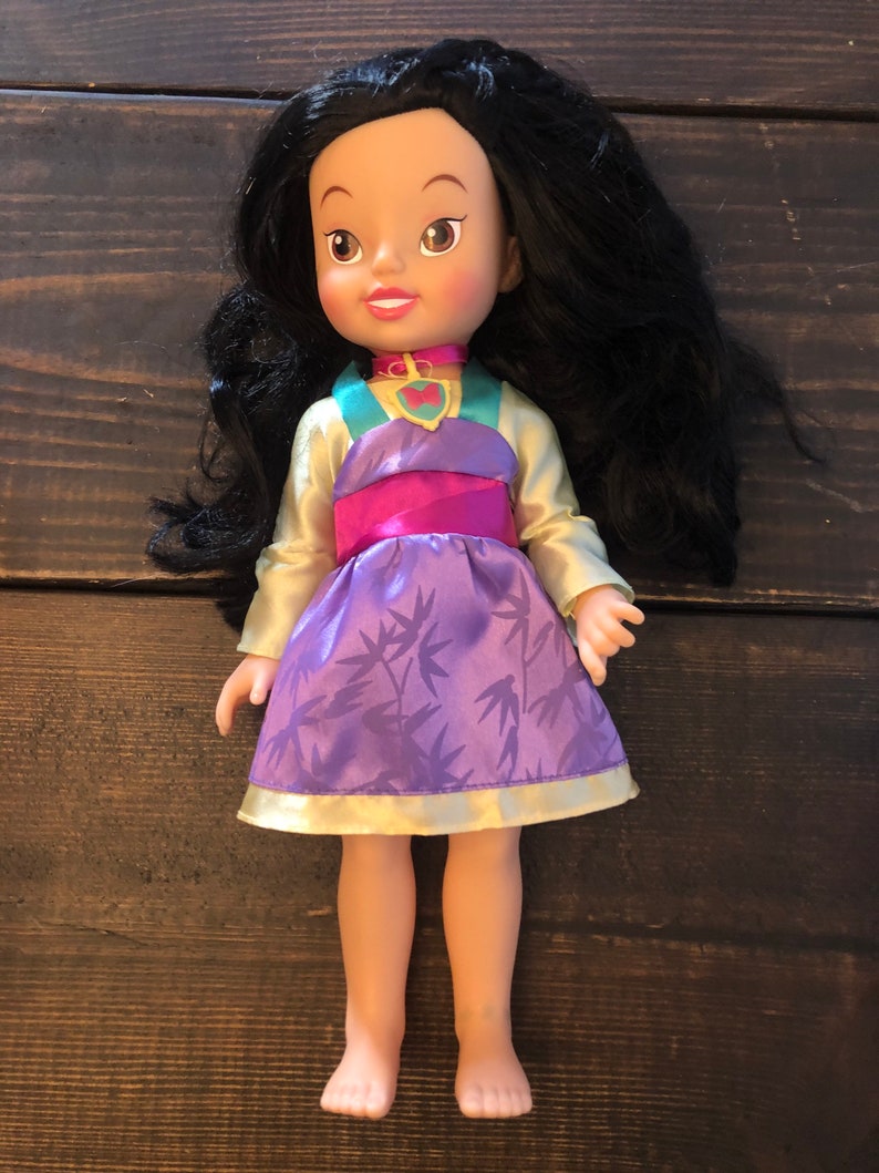 May include: A small doll with long black hair, wearing a purple and gold dress with a bamboo print. The doll has a pink and green top and a heart-shaped pendant necklace.