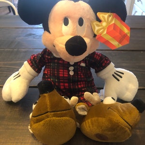 May include: A plush Mickey Mouse wearing red and black plaid pajamas and reindeer slippers. The plush toy is holding a gift box with a yellow bow.